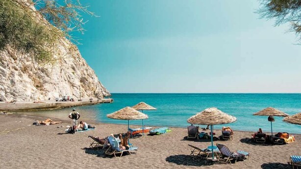 A beach with sun loungers, umbrellas, and a rocky cliff. - Spiridoula villa (Santorini)