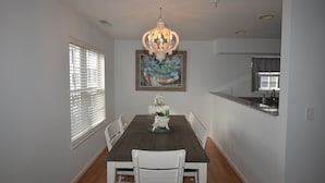 A dining area with a dark wooden table, white chairs, a chandelier, and a painting on the wall.