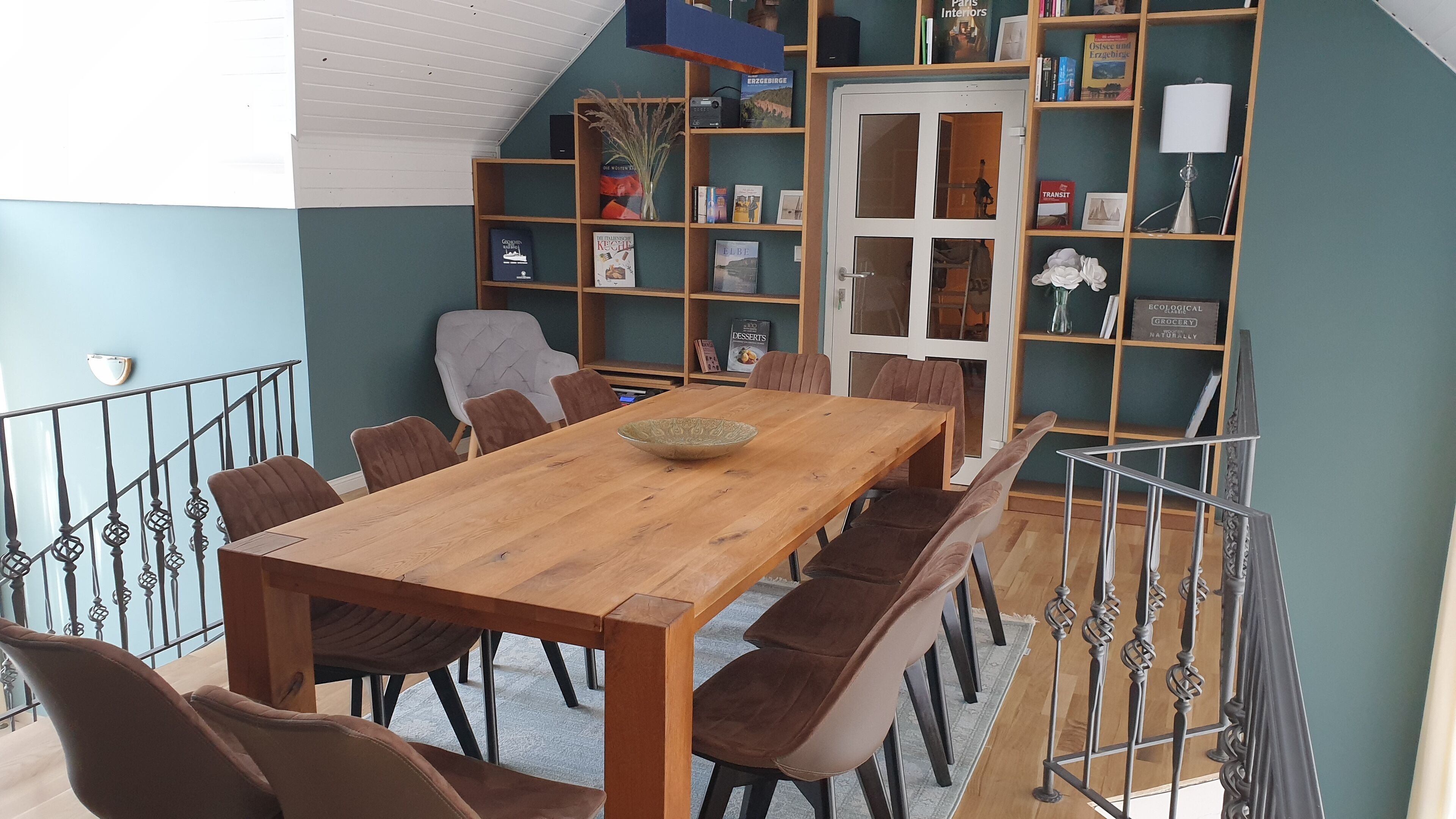 A dining area with a wooden table and brown upholstered chairs, a bookshelf with books and decorative items, and a staircase with a railing.