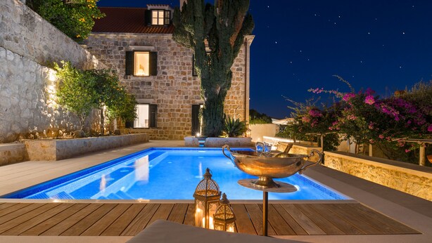Une piscine entourée d’un bâtiment en pierre en arrière-plan, d’une terrasse en bois et d’un ciel nocturne.
