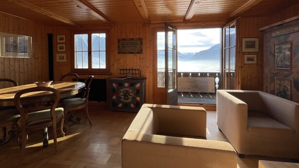 A wooden cabin interior with a dining area, a sofa, and a view of mountains through large windows. - Exclusive Chalet with amazing view (Benken)