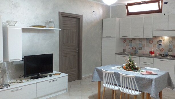 Una cucina compatta con mobili bianchi, un tavolo da pranzo con sedie, un televisore su un mobiletto bianco e una piccola ventola.