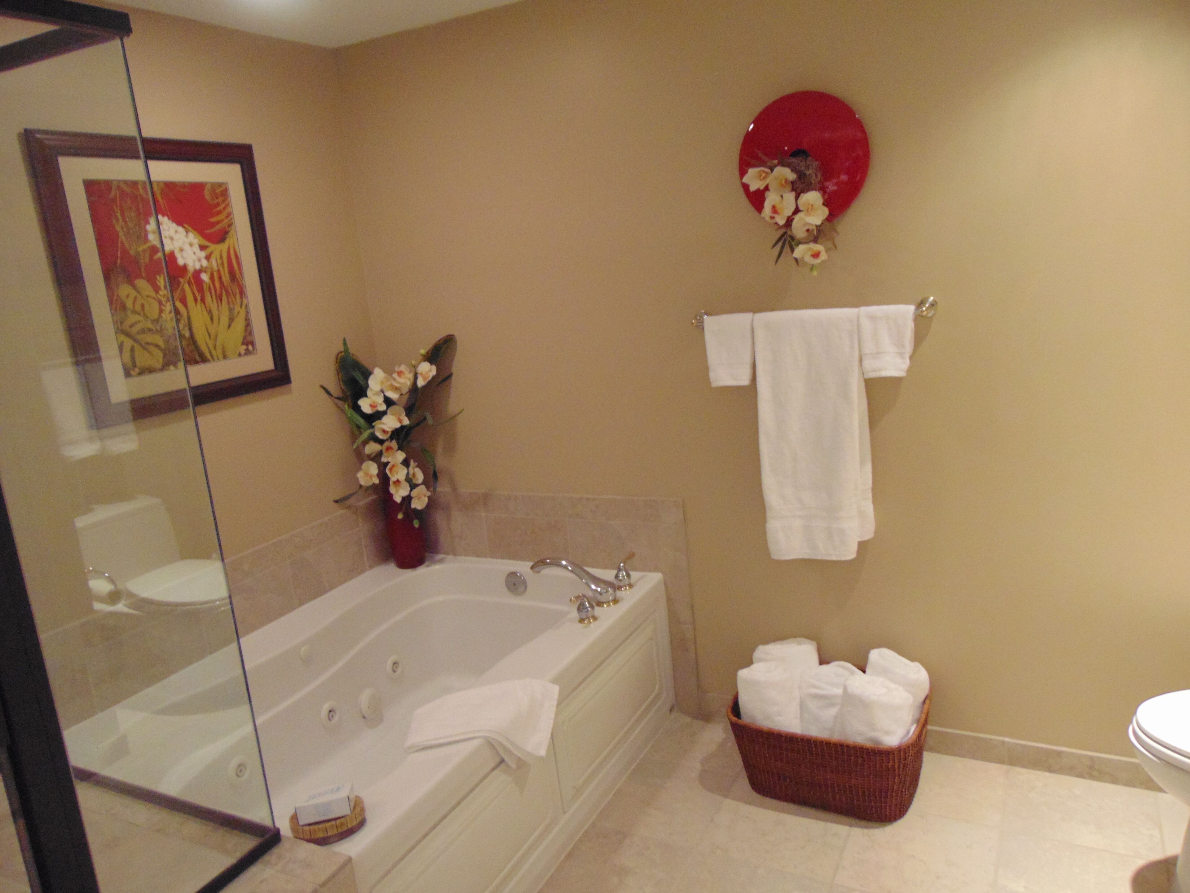 A bathroom with a bathtub, a glass shower enclosure, a sink, a toilet, a red decorative plate, a vase with flowers, and a basket of towels.