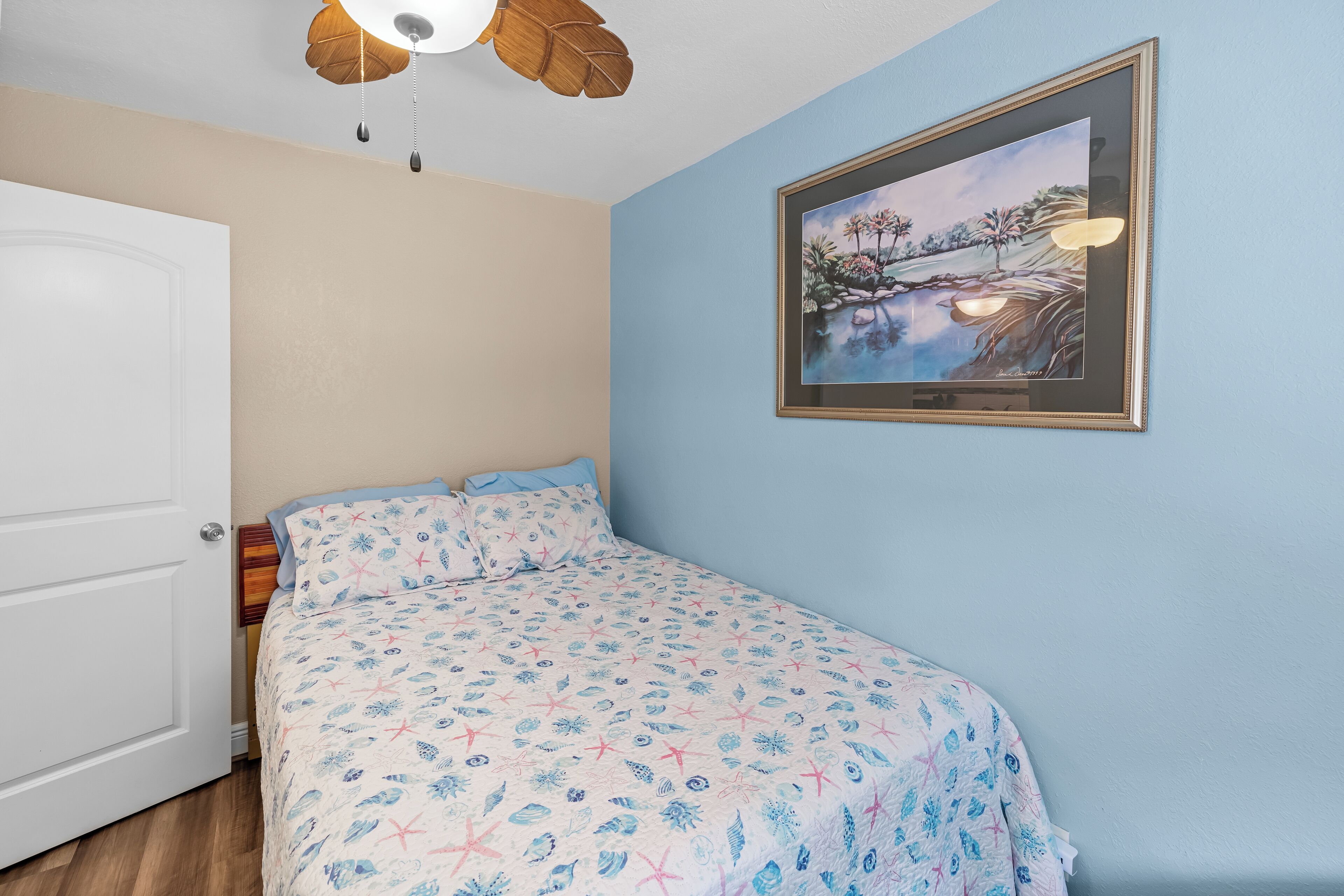 A bedroom with a bed, a door, a ceiling fan, and a framed painting on the wall.