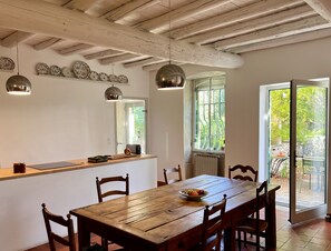 Un comedor con una mesa de madera, sillas, un frutero y una encimera con platos decorativos. Hay luces colgantes, una ventana con vistas y una puerta que da al exterior.