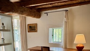 Una habitación rústica con una mesa de madera, dos sillas, una lámpara, un ventanal con cortinas y un estante con adornos.