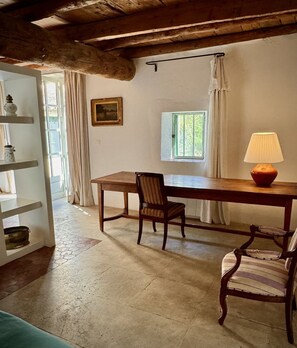 A rustic room with a wooden table, two chairs, a lamp, a window with curtains, and a shelf with decorative items.