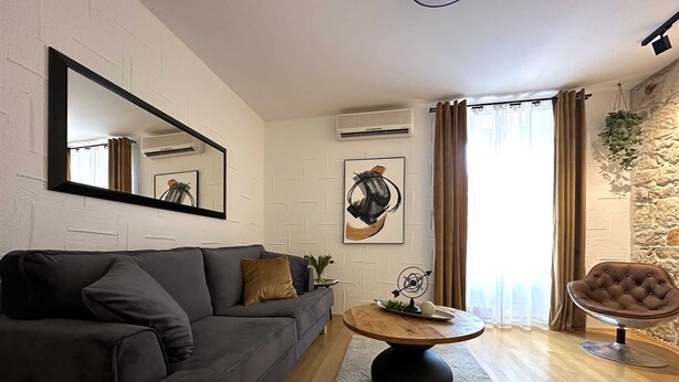 Un salon moderne avec un canapé gris foncé, une table basse ronde en bois et un fauteuil. Un grand miroir est accroché au mur, et un tableau abstrait encadré se trouve au-dessus du canapé.