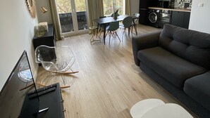 Ett modernt vardagsrum med en soffa, ett matbord och stolar. Här finns också ett köksområde med vitvaror och utsikt över utemiljön.