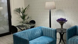 Een moderne woonkamer met een blauwe bank, een klein rond tafeltje met een vaas paarse bloemen, en een vloerlamp.