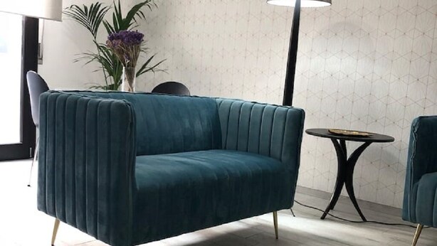 Een smaragdgroene fauteuil met een geweven bekleding, een klein rond tafeltje met een zwarte voet en een hoge vloerlamp met een witte lampenkap.