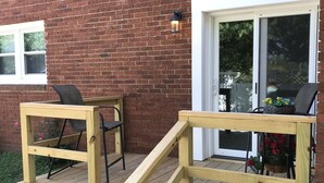 Een houten terras met een helling naar een deur, een stoel en een bloempot.