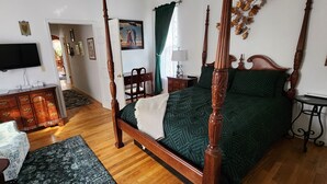 A four-poster bed with a green bedspread, a wooden nightstand with a lamp, a television mounted on the wall, and a wooden dresser with drawers. - Happy Days and Nights - Valiant (Harbor Springs)