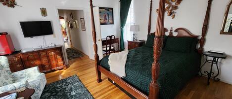 A four-poster bed with a green bedspread, a wooden nightstand with a lamp, a television mounted on the wall, and a wooden dresser with drawers.