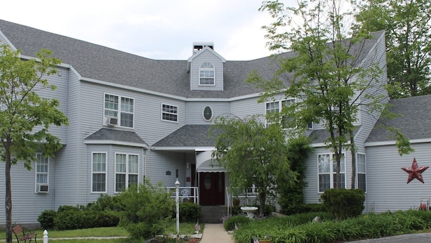 A large, grey, two-story house with a symmetrical design, featuring a central entrance with a covered porch, multiple windows, and decorative red stars on the facade. - Happy Days and Nights - Valiant (Harbor Springs)