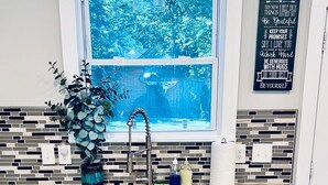 Una cocina con armarios blancos, un lavamanos de acero inoxidable, una ventana con vistas a árboles y un cartel en la pared que dice 'Family Rules'.