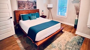 Un dormitorio con cama, ventilador de techo, pintura de paisaje urbano, lámpara de pie, ventana con persianas y un cartel motivacional.