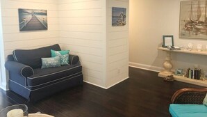 Un salon doté d’un parquet en bois foncé, d’un canapé noir agrémenté de coussins turquoise et d’un mur blanc orné de lignes horizontales. On y trouve deux tableaux encadrés, une petite table avec des livres et des objets décoratifs, ainsi qu’une chaise en osier avec un coussin turquoise.