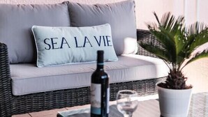 Un sofà de vimetera amb un coixí que diu "SEA LA VIE" i una ampolla de vi sobre una taula a prop.