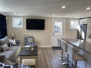Una sala de estar moderna con un sofá, una mesa de centro y un televisor. Hay una zona de cocina con un refrigerador y taburetes de bar.