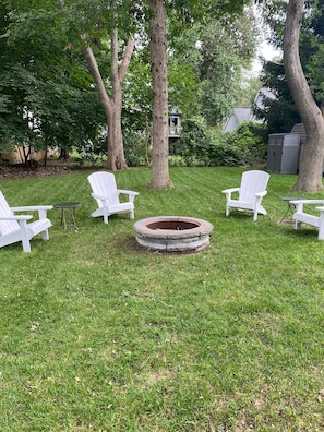 Relax in a serene backyard oasis with a fire pit and Adirondack chairs surrounded by lush greenery.