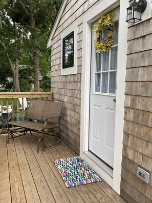 Une terrasse en bois avec une chaise et un tapis coloré, une porte blanche ornée d’une guirlande, et un tableau encadré de noir au-dessus de la porte.