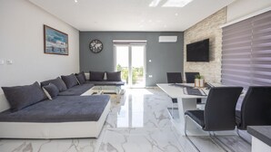 Ein modernes Wohnzimmer mit einer Eckcouch, einem Esstisch mit Stühlen, einem an der Wand befestigten Flachbildfernseher und einem großen Fenster mit Jalousien.