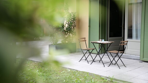Une terrasse extérieure avec une table et des chaises, un pot de fleurs et un bâtiment aux volets verts.