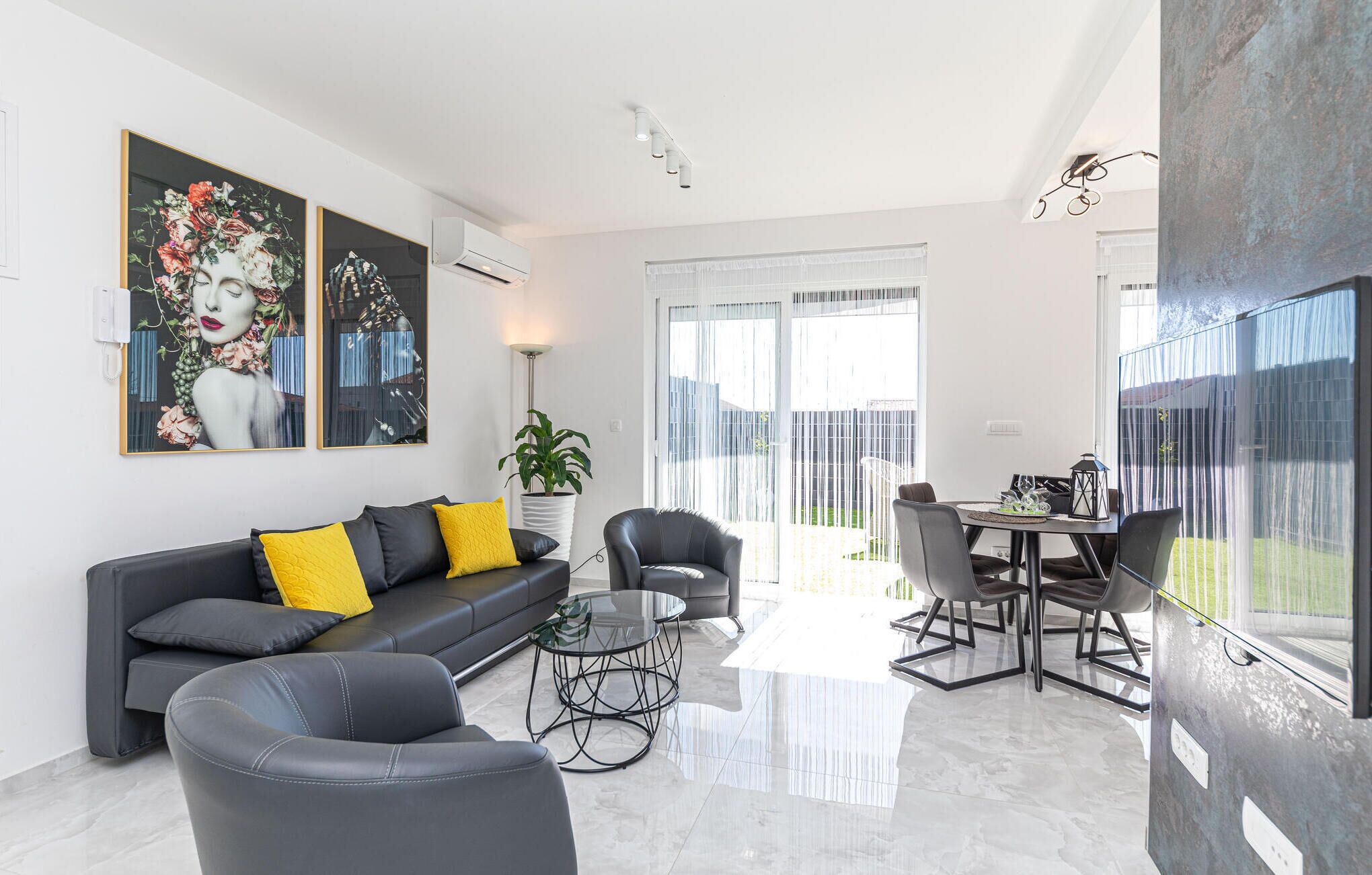 A modern living room with a black leather sofa, a glass coffee table, and a dining area with a table and chairs. There are two large paintings on the wall, a potted plant, and a sliding glass door leading to a balcony.