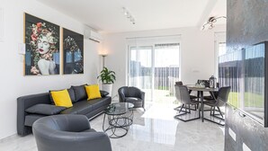 Una sala moderna con un sofá de cuero negro, una mesa de centro de vidrio y un área de comedor con una mesa y sillas. Hay dos cuadros grandes en la pared, una planta en maceta y una puerta corrediza de vidrio que da a un balcón.