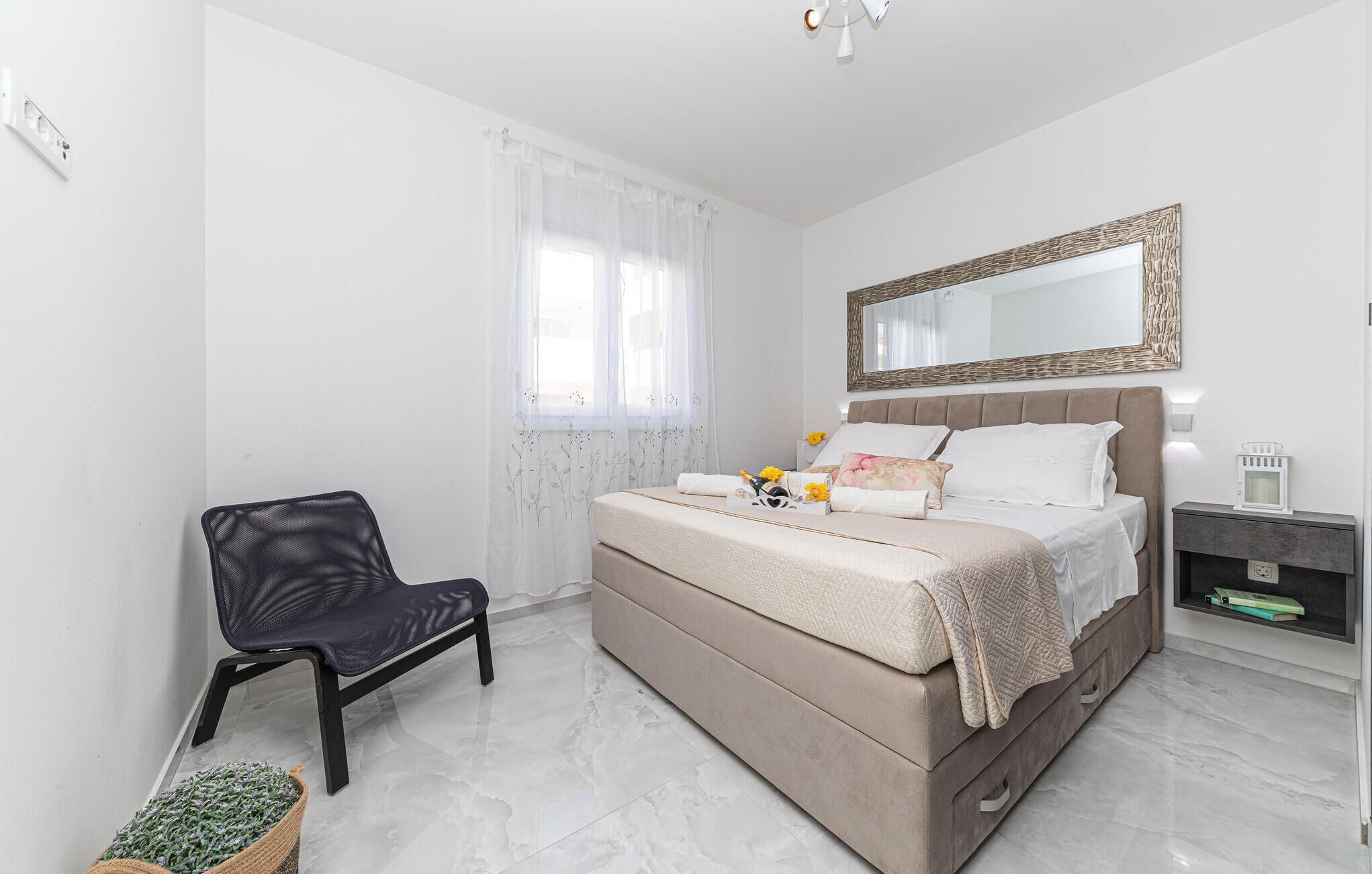 A bedroom with a bed, a chair, a bedside table, a mirror, and a window with sheer curtains.