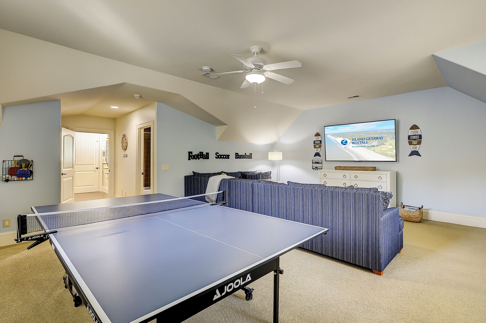 A room with a ping pong table, a ceiling fan, a TV displaying an island getaway rental ad, and a wall with sports-themed decorations.
