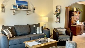 Een woonkamer met een grijze bank, een houten salontafel, een klein voetenbankje en een lamp.