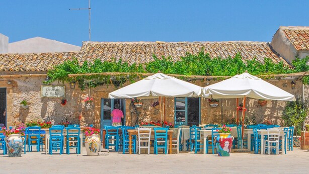 An outdoor café with tables and chairs, a stone building with a tiled roof, and greenery. - Gorgeous home in Rosolini (Rosolini)