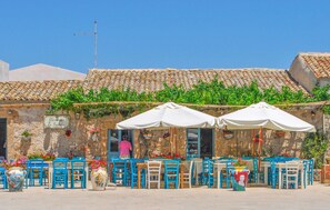 An outdoor café with tables and chairs, a stone building with a tiled roof, and greenery. - Gorgeous home in Rosolini (Rosolini)