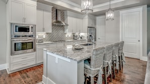 A modern kitchen with white cabinetry, stainless steel appliances, a brick backsplash, and a central island with bar stools. - Luxury North Beach Resort home w/ private pool, master on main & open floor plan (North Myrtle Beach)