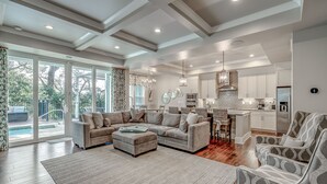 A spacious living room with a sectional sofa, ottoman, and patterned armchairs. A large window offers a view of a pool and outdoor area. - Luxury North Beach Resort home w/ private pool, master on main & open floor plan (North Myrtle Beach)