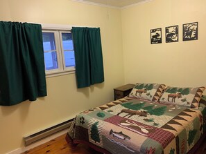A bedroom with a bed, a window with curtains, a wall with framed pictures, and a heater on the floor. - Hide-A-Way Cabin, Pool & AC, Authetic Adirondack Cabin Steps from Loon Lake (Chestertown)