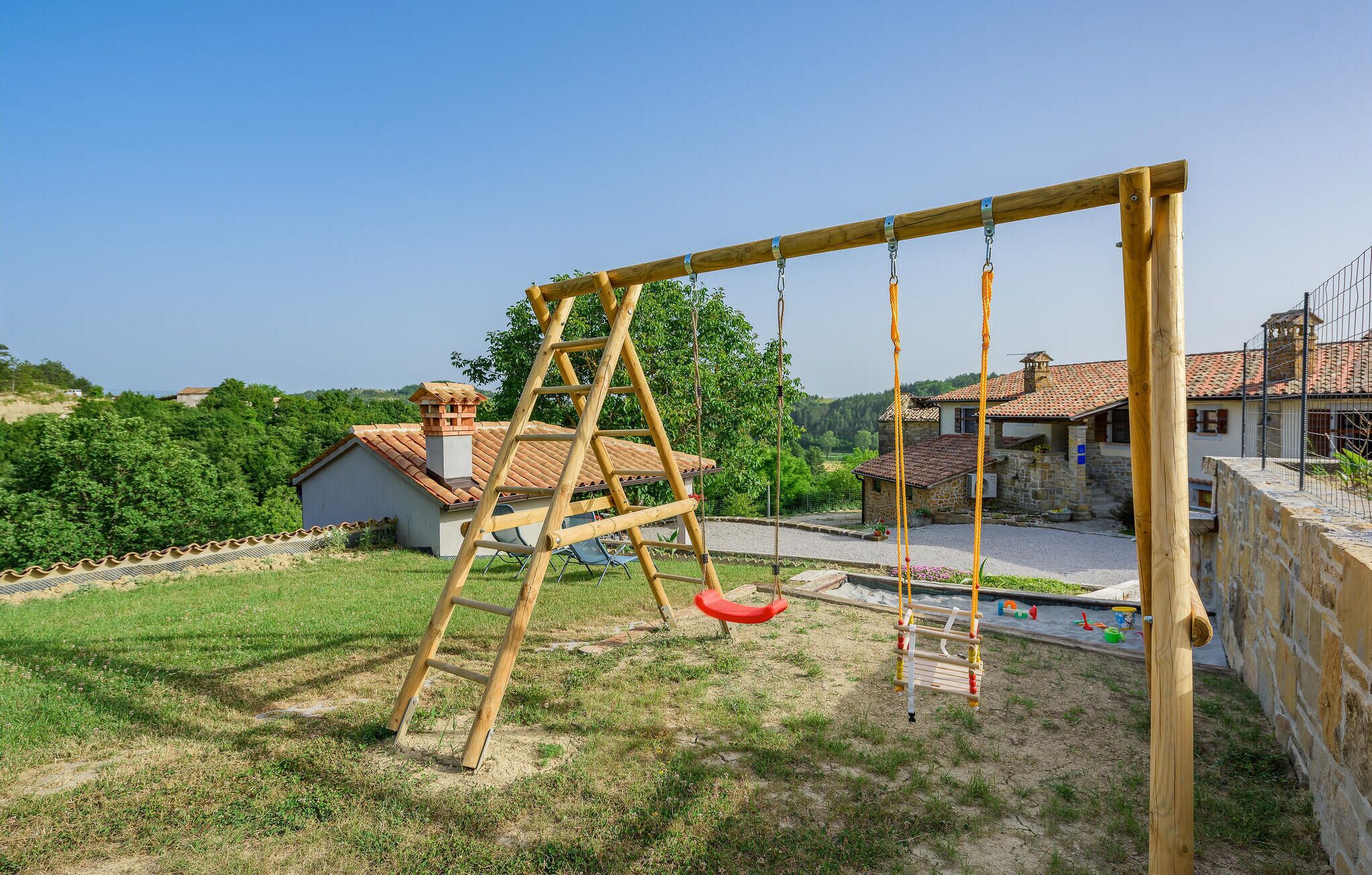 Een houten schommel in een gazon met een stenen muur en een huis met pannendak op de achtergrond.