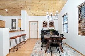 Un espace repas doté d’un plafond en bois, d’un lustre, d’une table à manger avec des chaises, d’un bar avec des tabourets et d’un coin cuisine avec des placards et une fenêtre.