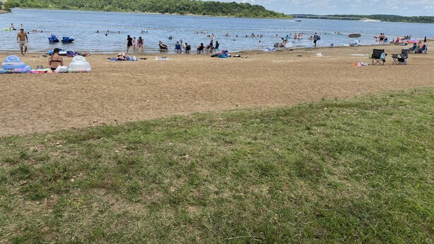 Una playa con gente nadando y tomando el sol, un área verde al frente y un lago al fondo.