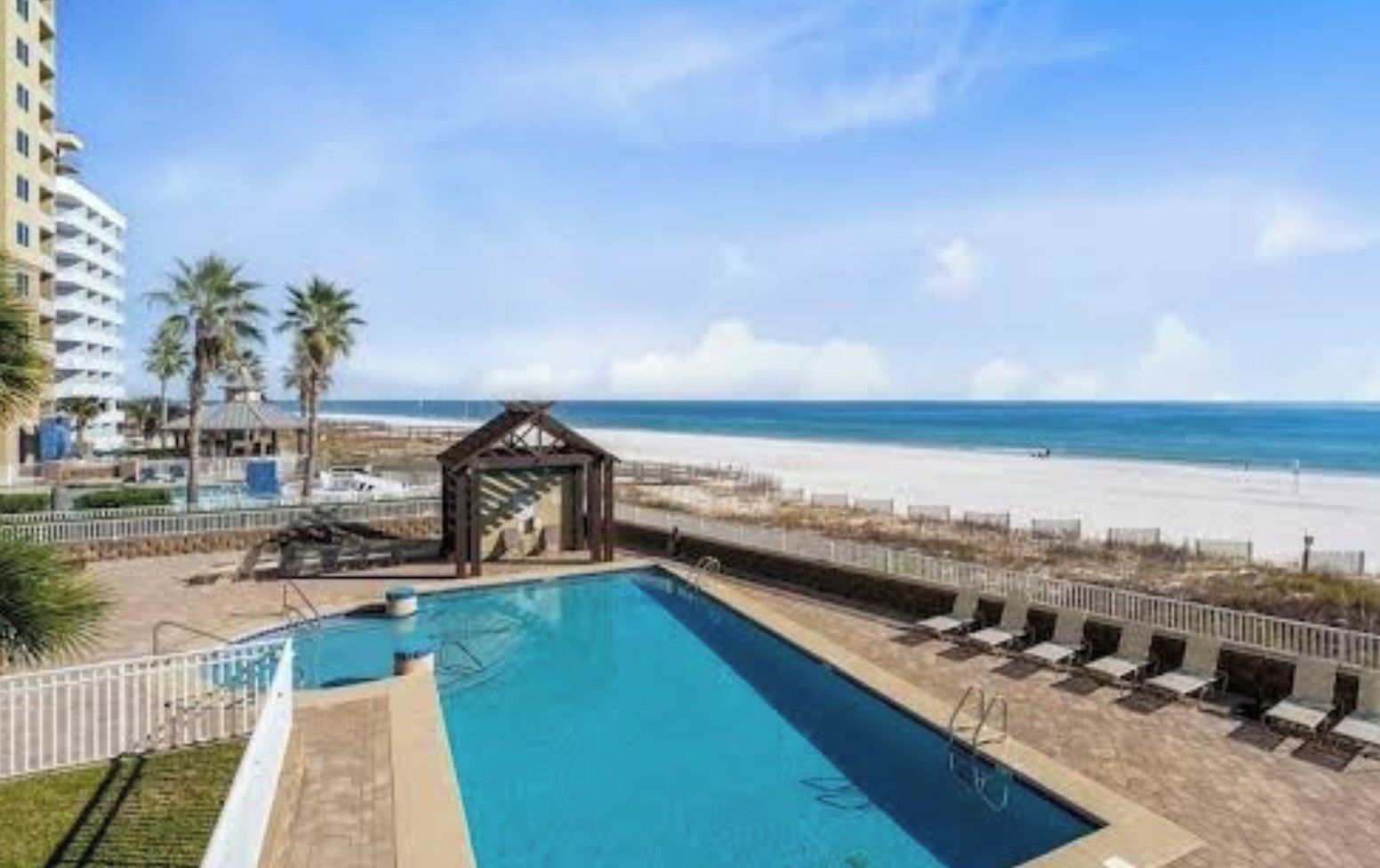 A swimming pool with a beach view, a covered seating area, and a tall building in the background.
