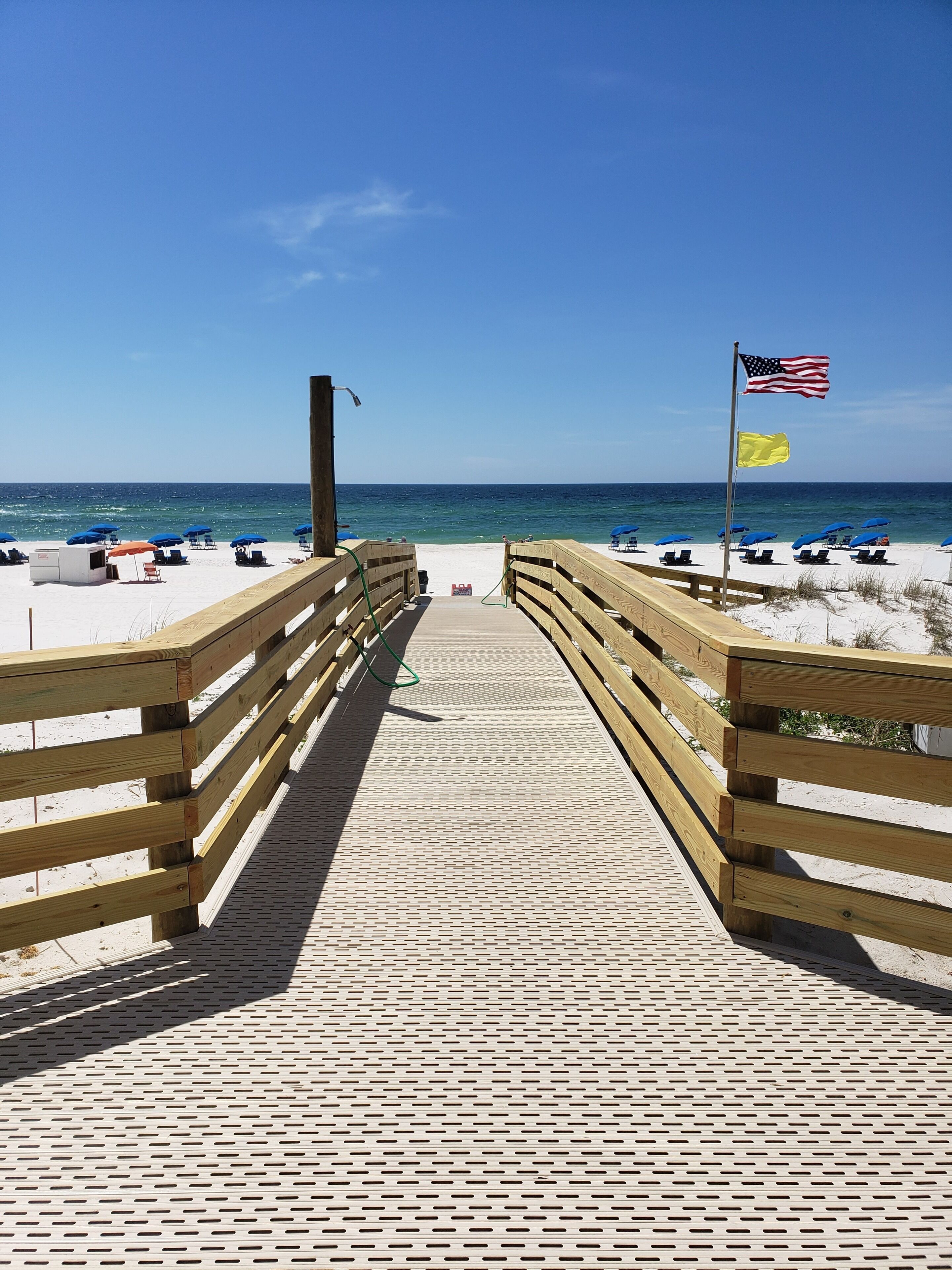 A wooden boardwalk leading to a beach with blue umbrellas and a flag.