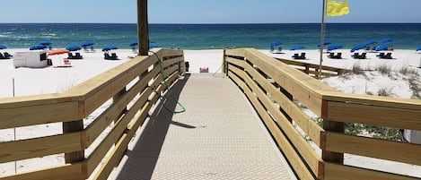 Ein hölzerner Steg führt zu einem Strand mit blauen Sonnenschirmen und einer Flagge.