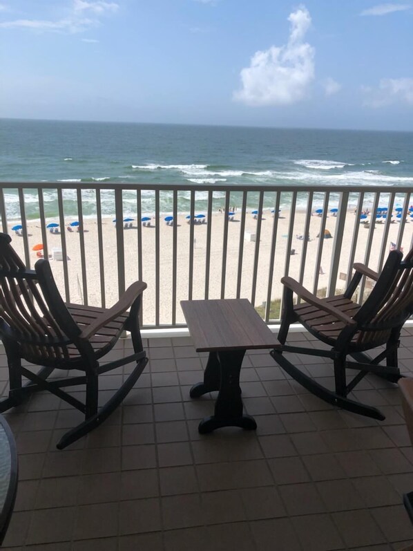 A balcony with two rocking chairs and a small table, overlooking a beach with umbrellas and people. - 6th Floor Gulf Front Corner - 3 bedrooms, 3 baths (Orange Beach)