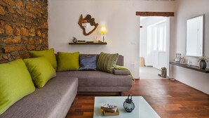 A cozy living room with a stone wall, a wooden ceiling, a grey sofa with green and blue cushions, a small table with a lantern, and a wall-mounted shelf with a lamp and decorative items. - Amazing home in Barban with sauna (Barban)