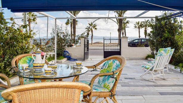 Une terrasse extérieure aménagée avec des meubles en osier, une table en verre et une vue sur la plage et les montagnes.