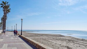 Une promenade dallée, bordée d’un muret en pierre et de palmiers, qui mène à une plage de sable.