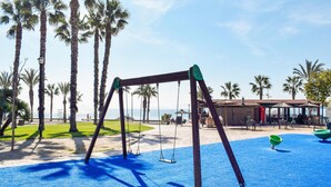 Un terrain de jeux avec des balançoires et une aire de jeux bleue, entouré de palmiers et offrant une vue sur la plage.