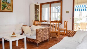 Un salon avec un canapé blanc, un fauteuil en osier, une table à manger avec des chaises et une fenêtre avec des stores.